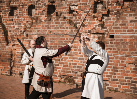 Dwóch mężczyzn w strojach zakonnych walczących mieczami na tle zamku.