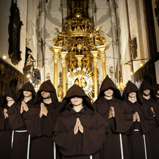 9 osób w strojach mnichów z założonymi kapturami składającymi ręce do modlitwy w kościele.