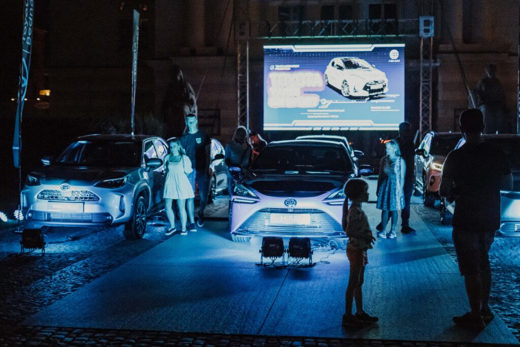 Na zdjęciu grupa ośmiu ludzi chodzi i ogląda 6 samochodów marki Toyota