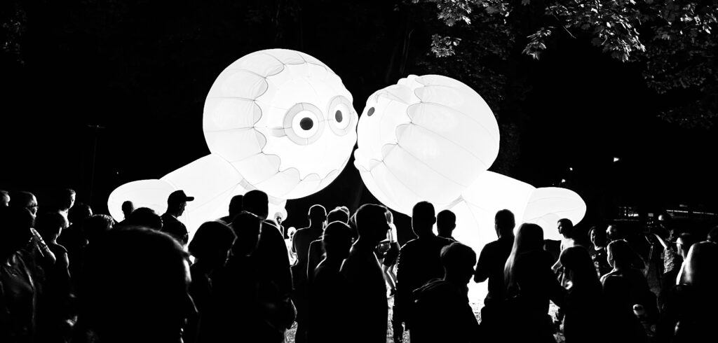 Na zdjęciu znajduje się tłum oglądający dwa białe, duże balonowe, postacie przypominające wyglądem eskomosy i dające sobie całusa w policzek