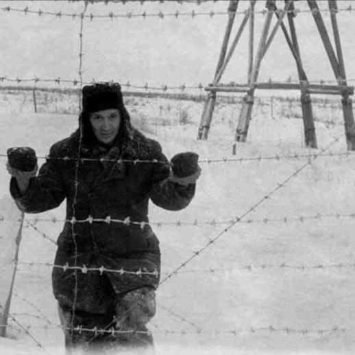 Grafika przedstawia ciepło ubranego mężczyznę znajdującego się za drutem kolczastym w niewoli na Syberii, tło jest czarno- białe