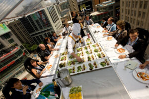 Na zdjęciu widać platformę w formie eleganckiego stołu restauracyjnego. Ludzie jedzą. Jest też kucharz serwujący posiłki.