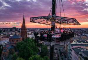 Zdjęcie z logo dinner in the sky. Na zdjęciu widać platformę w formie stołu unoszoną przez dźwig, na tle miasta.