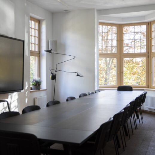 Wnętrze Kulturalnego hubu Bydgoskiego Przedmieścia. Sala konferencyjna, na środku której stoi długi stół, a wokół niego krzesła. Po lewej stronie wisi duży telewizor, obok lampa stojąca. Po prawej stronie duże okno z widokiem na park.