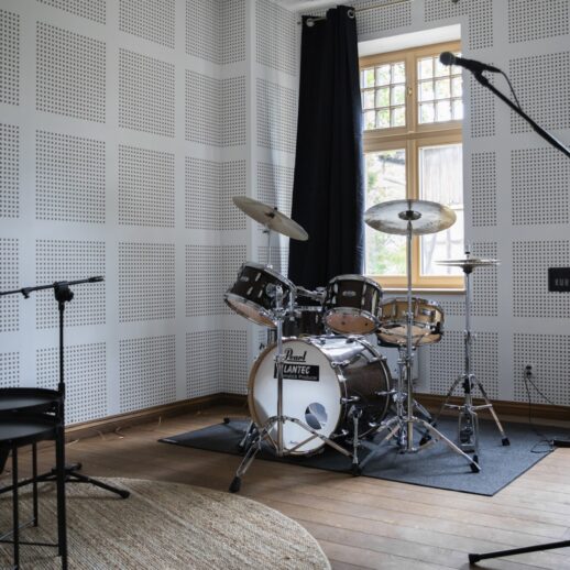 Wnętrze Kulturalnego hubu Bydgoskiego Przedmieścia. Studio nagrań, w którym stoi perkusja, mikrofon na statywie i keyboard. Na podłodze dwa dywany - czarny i beżowy.