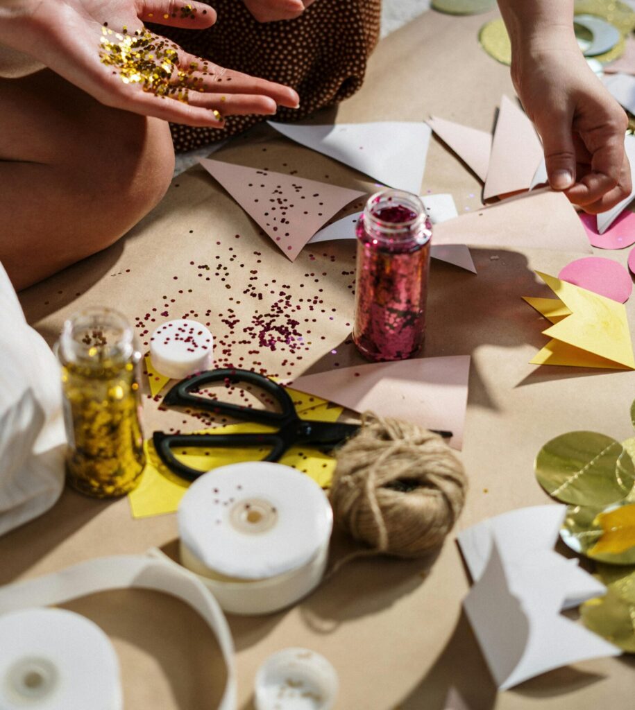 Rozrzucone materiały do robótek ręcznych. Kartki papieru, origami, nożyce, fiolki z brokatem, kłębek nitki, taśmy.