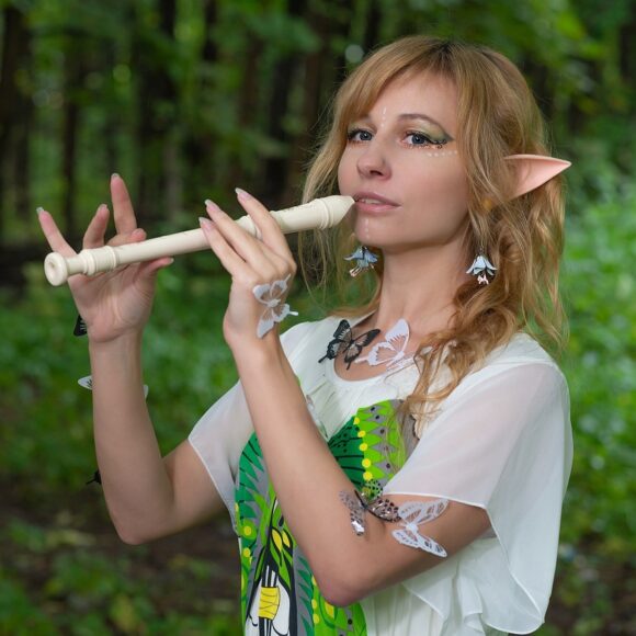Blondwłosa dziewczyna z długimi uszami, jak u elf. Ma przyklejone do skóry białe i czarne motylki. Stoi w lesie i gra na flecie.