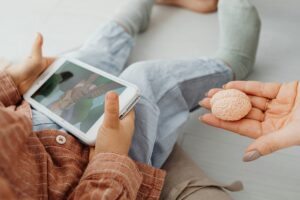 Zdjęcie przedstawiające małe dziecko oglądające bajkę na telefonie. Obok dłoń dorosłej osoby trzymająca figurkę mózgu.