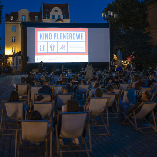 Zdjęcie przedstawiające publiczność oglądającą film w trakcie wydarzenia Kino plenerowe. Na ekranie plakat wydarzenia.